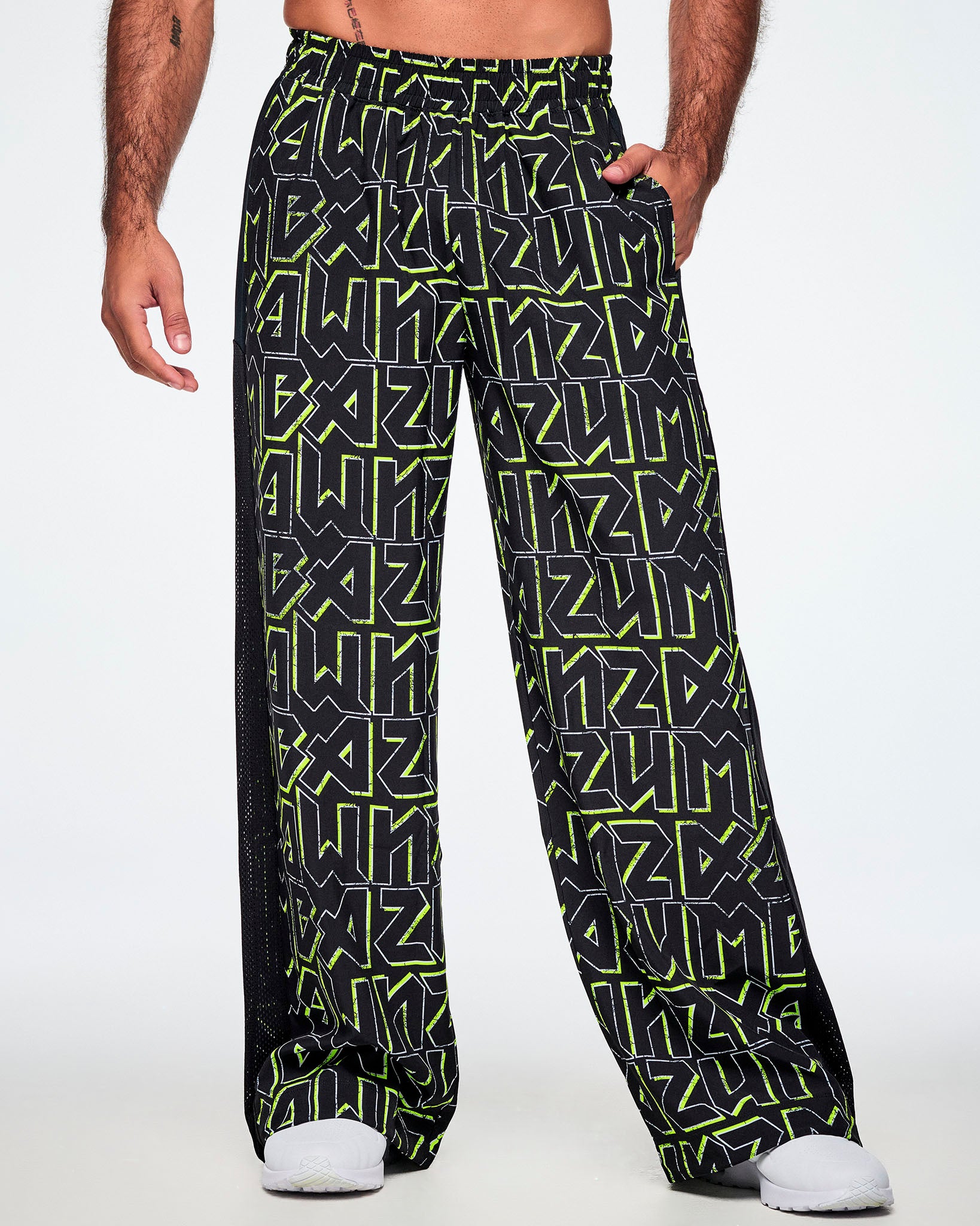 A male model shown from the front, with a hand in his pocket, wearing wide leg track pants, with ZUMBA text in an all over black pattern outlined in Zumba green colors with a black mesh side panel strip and paired with white shoes.