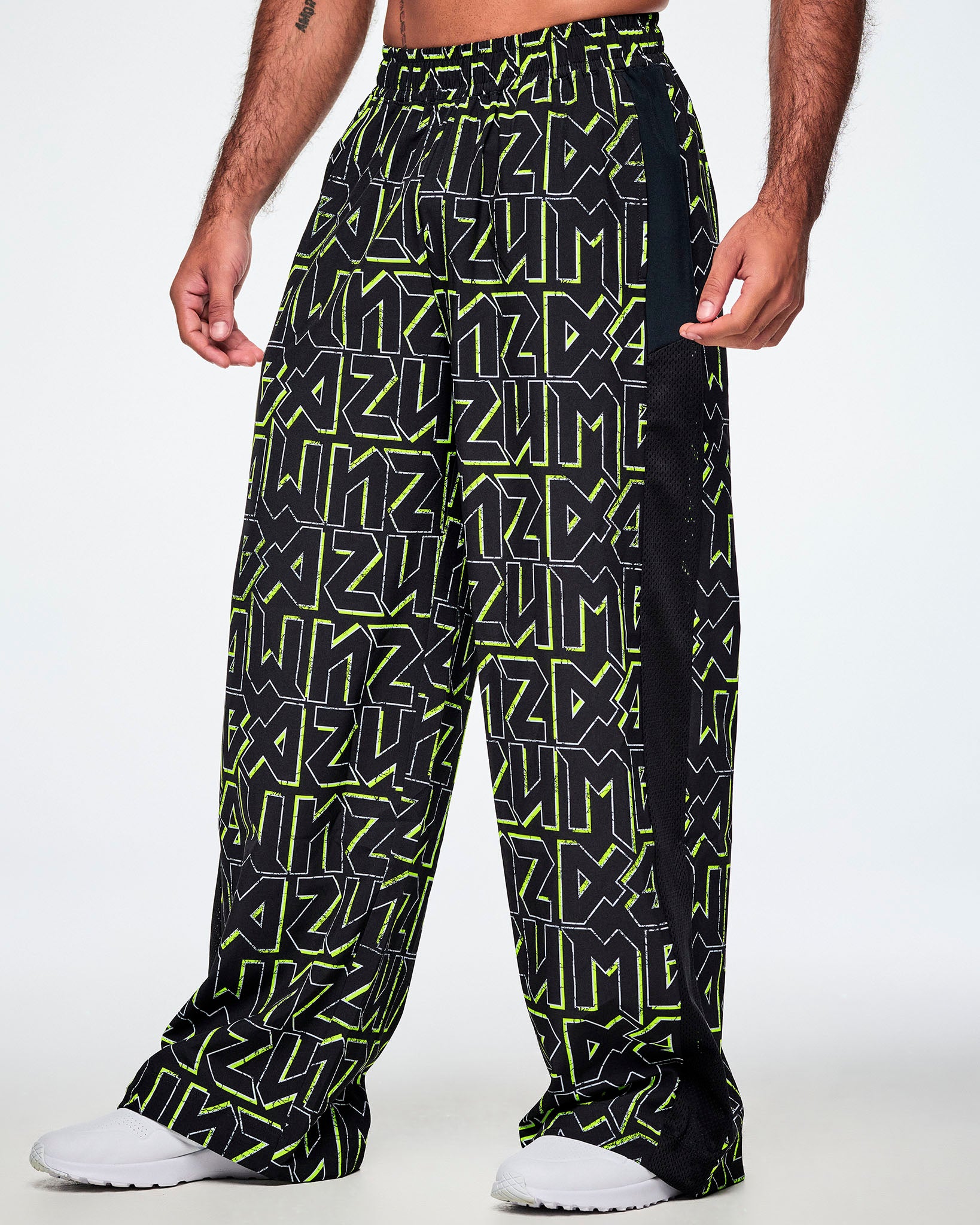 A male model wearing wide leg track pants, with ZUMBA text in an all over black pattern outlined in Zumba green colors with a black mesh side panel strip and paired with white shoes.