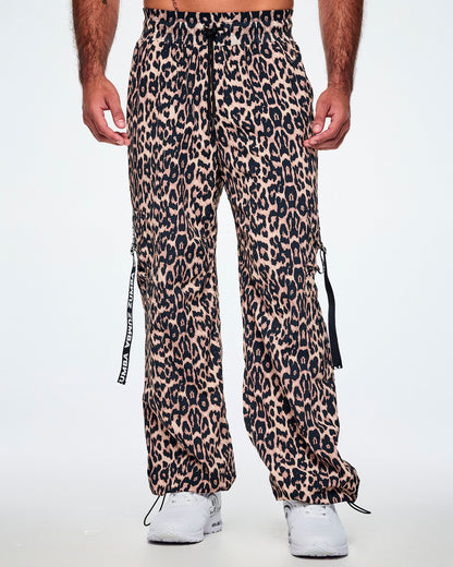 Front view of a male model wearing tan leopard print cargo pants with black Zumba straps on the pockets, paired with white shoes.