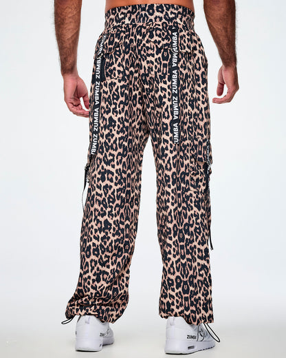Back view of a male model wearing tan leopard print cargo pants with black Zumba straps on the pockets, paired with white shoes.
