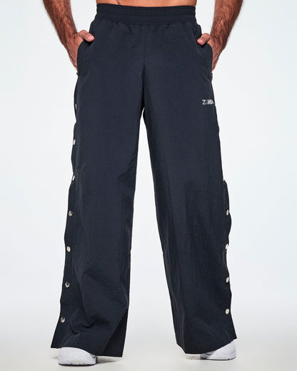 Front view of a male model with both hands in his pockets, wearing wide-leg black pants with a silver 'ZUMBA' logo on the left thigh and decorative silver-tone snap buttons down the outer side of the legs, shown against a white background.