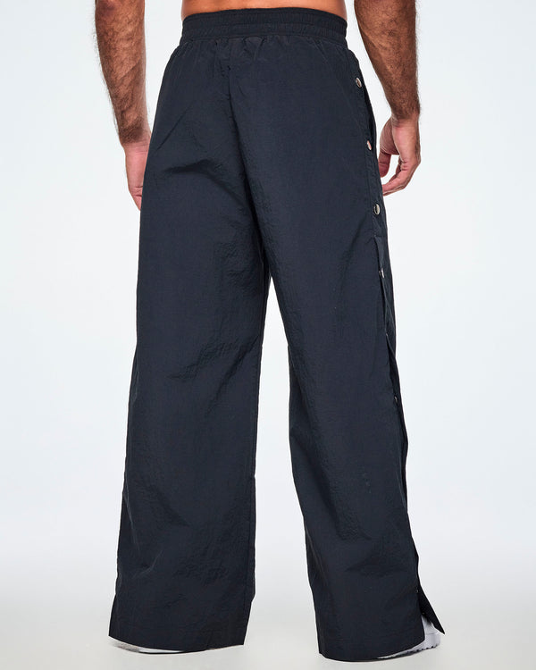 Back view of a male model wearing wide-leg black pants with silver-tone snap buttons down the outer side of the legs, shown against a white background.