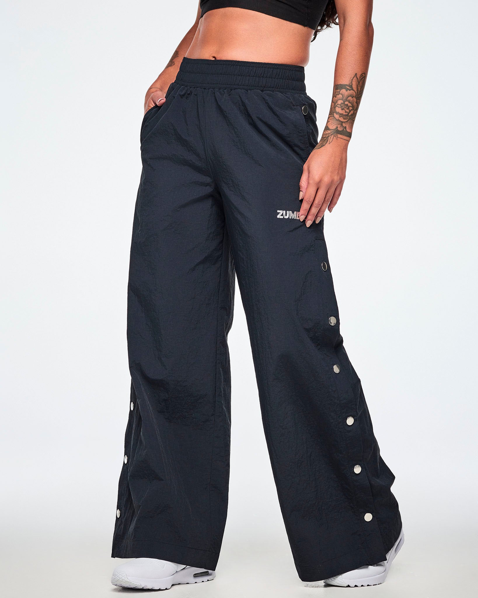 A female model wearing wide-leg black pants with a silver 'ZUMBA' logo on the left thigh and decorative silver-tone snap buttons down the outer side of the legs, shown against a white background.