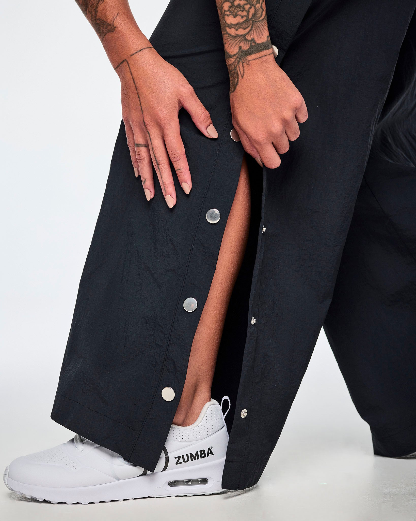 A close-up of a female model wearing black wide-leg pants showing off the side snaps being unfastened at the lower leg and paired with a white and black Zumba shoe.