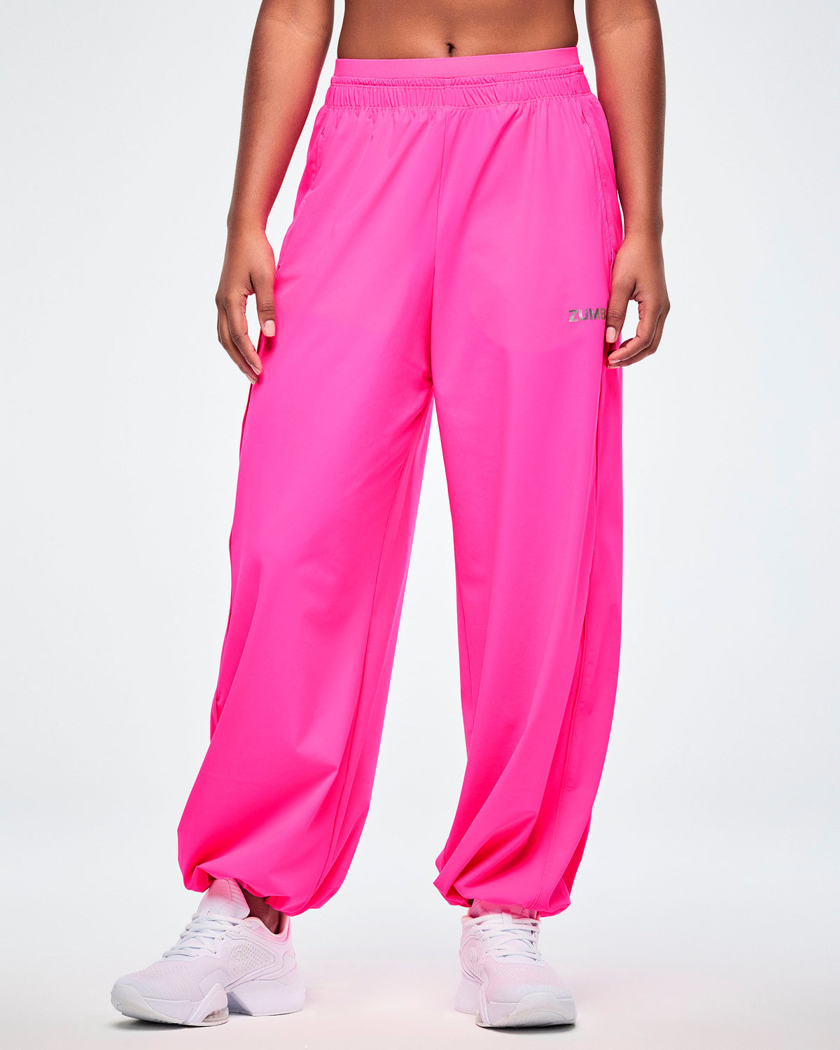 Pink slouch pants with a small silver 'ZUMBA' text on the left leg, worn by a female model, paired with white shoes.