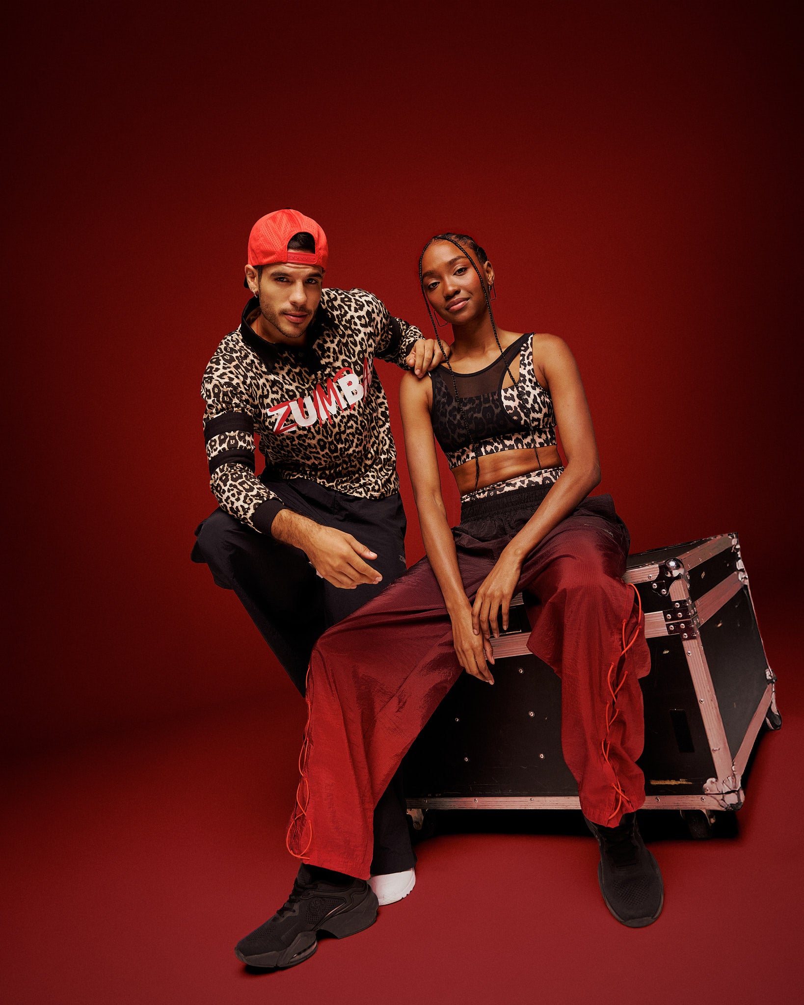 A male and a female model pose together in a studio setting with a solid dark red background and sitting on an equipment case. The man wears a red backwards hat, a leopard-print long-sleeve shirt with 'ZUMBA' printed on the front, black pants, and white sneakers. The female wears a leopard print bra with asymmetrical mesh overlay and black and red gradient pants with black shoes.