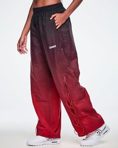 A female model wearing loose, wide-leg gradient pants that fade from black at the top to deep red at the bottom and showing the lower leg panel with adjustable criss-cross lace up detail and paired with white shoes.