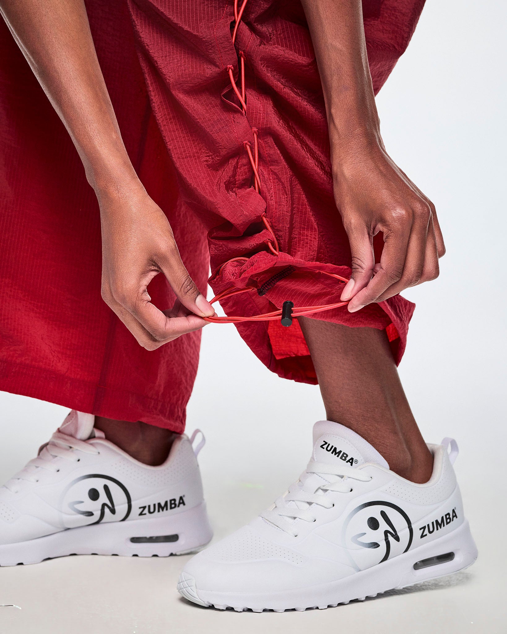A close-up view of a female model wearing black and red gradient pants and pulling on the adjustable criss-cross lace up detail from the ankles and paired with white Zumba shoes.