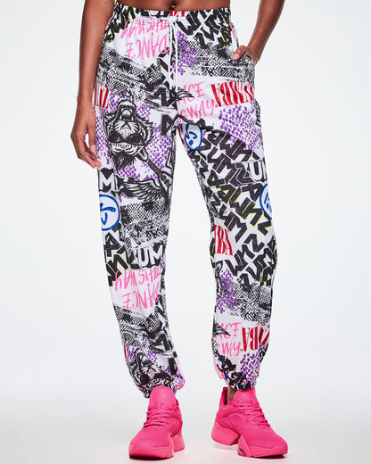 Front view of a female model, with a hand in her pocket of the white woven pants with an all-over ripped newspaper like pattern with black, white and red ZUMBA text and pink Dance this way text along with other Zumba rock graphics, Zumbitos and purple and black snakeskin patterns, paired with pink shoes.