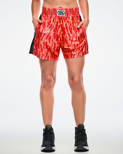 Front view of a female model with both hands in her pockets, wearing orange boxing shorts with an allover red “ZUMBA” monogram, paired with black-and-white shoes.