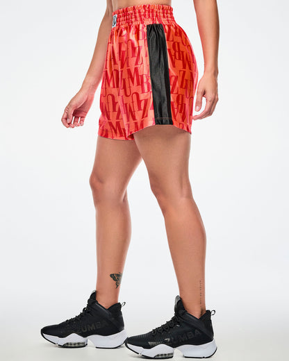 Side view of a female model wearing orange boxing shorts with an allover red “ZUMBA” monogram, paired with black-and-white shoes.