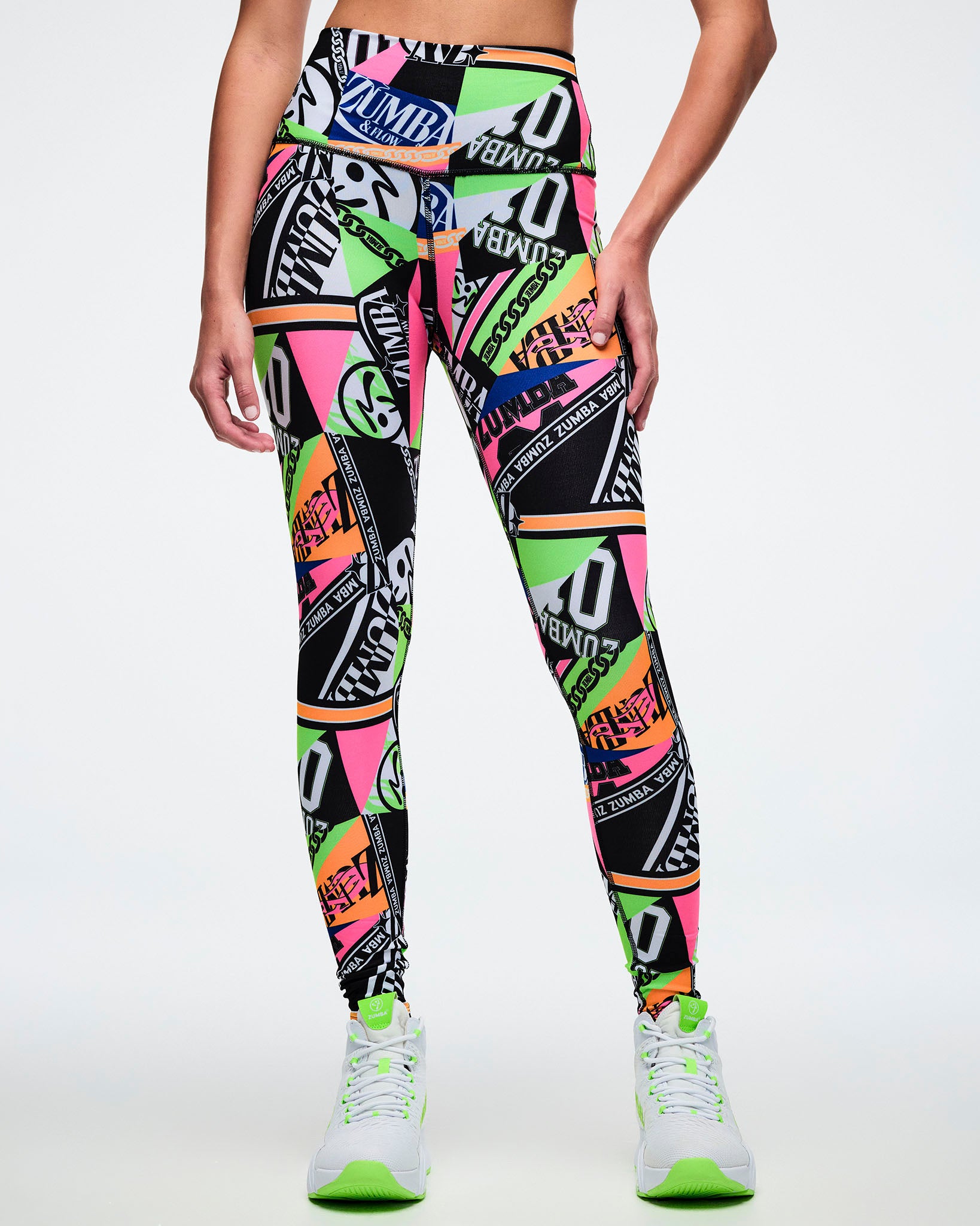 Front view of female model wearing multi-colored Zumba leggings with a geometric pattern paired with matching green and white shoes.