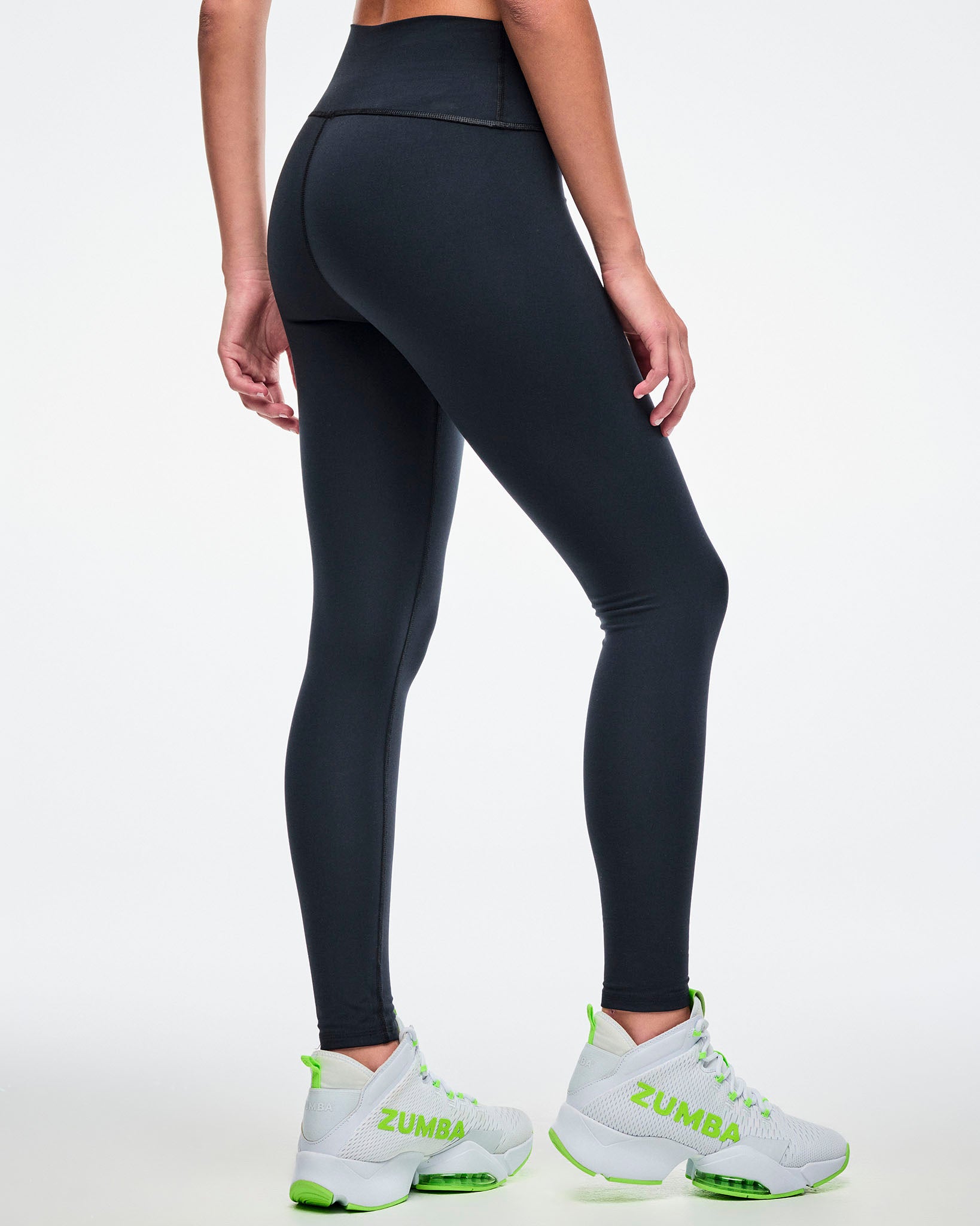 Back view of a female model wearing black Zumba leggings paired with green-and-white shoes.