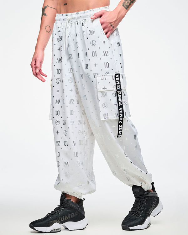 Male model in white cargo pants printed with silver Zumba motifs (ZW, Zumbito, dots, stars, “01”), featuring a black‑and‑white ribbon on the cargo pocket reading “Zumba,” in white, paired with black‑and‑white shoes.