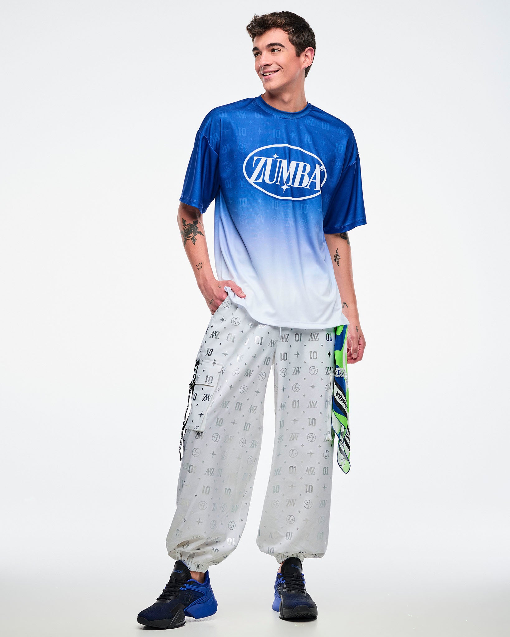 Male model in a blue and white gradient top with white cargo pants printed with silver Zumba motifs and paired with black and white shoes.