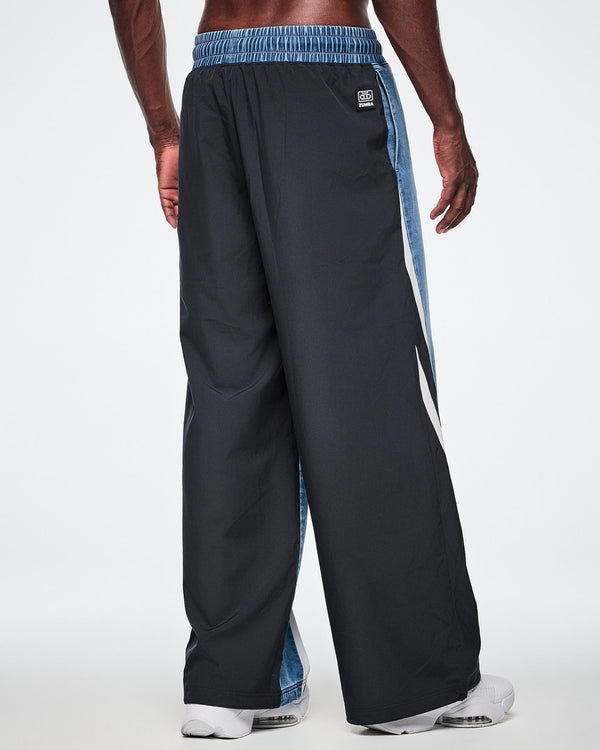 Back view of a male model wearing denim combo pants, showing the solid black back with a denim strip visible along the inseam and a Zumba cassette tag at the waistband.
