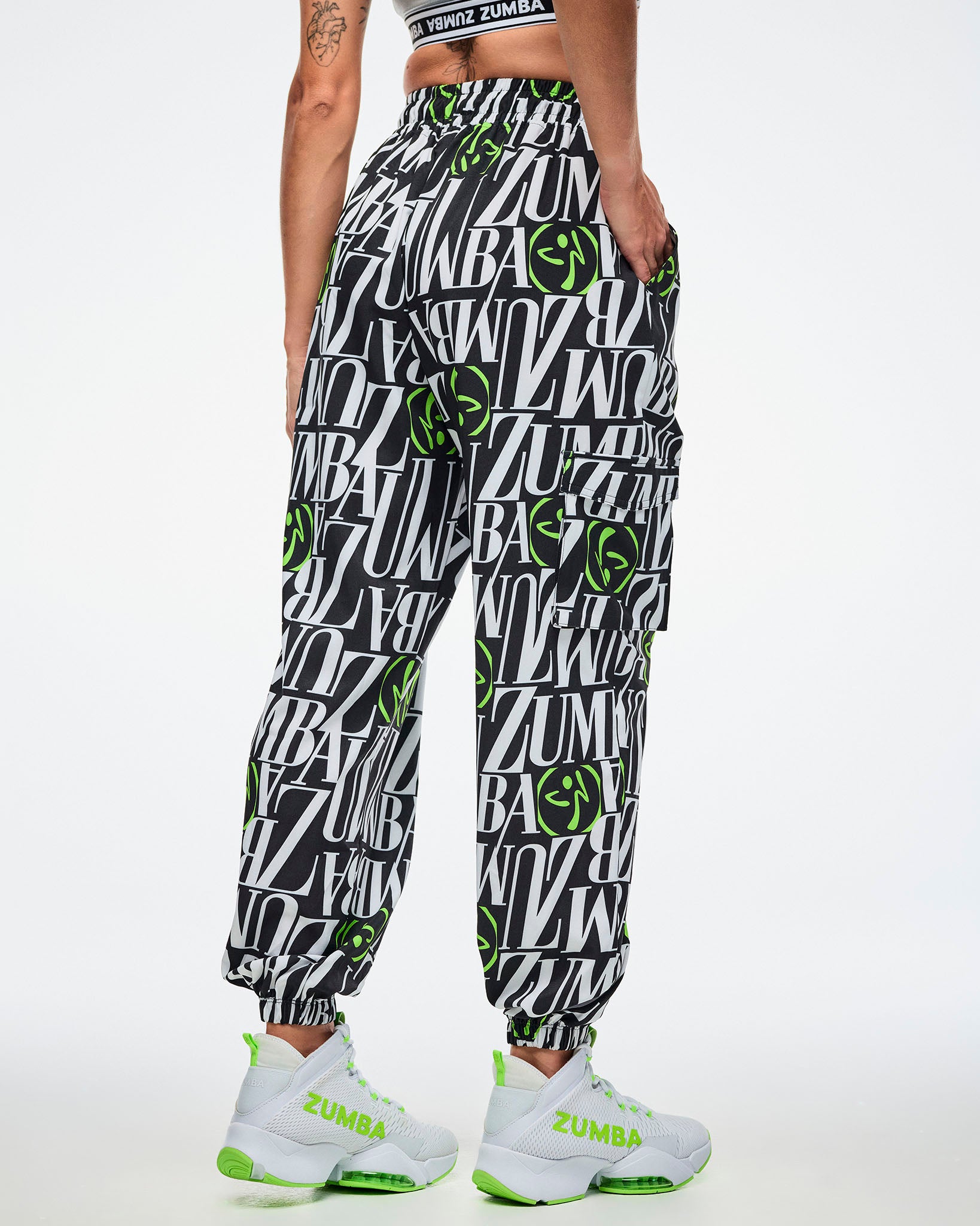 Back view of a female model wearing black and white patterned ZUMBA pants with green Zumbito accents, paired with white and green sneakers on a white background.
