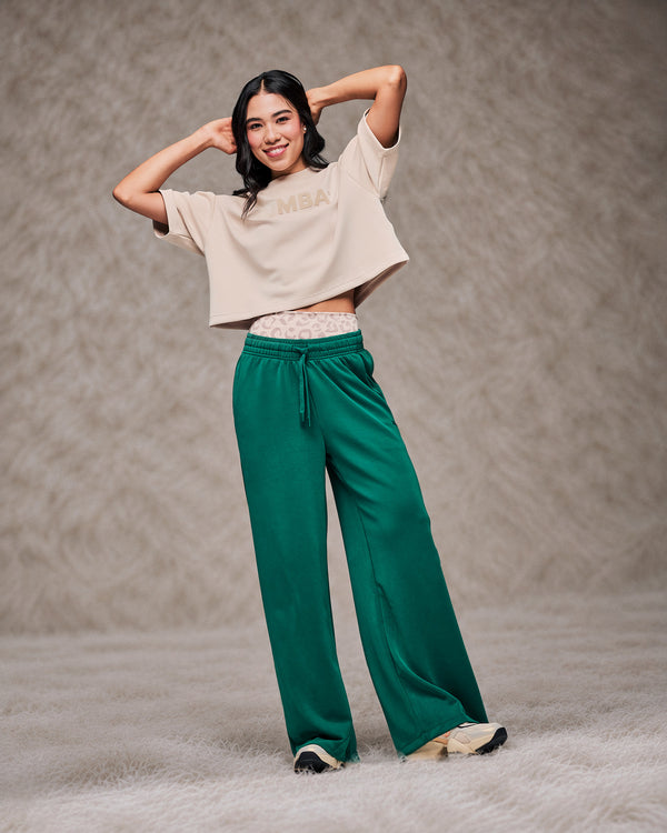 Female model with both hands behind her head in the teal green high-waisted pants, styled with a beige drop shoulder crop top. Shot on a shaggy cream rug against a taupe faux fur backdrop.