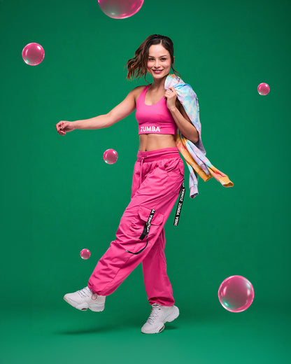 Female model in pink Zumba racerback and matching cargo pants with a tie-dye hoodie in her hand dancing on a green background with pink bubbles.