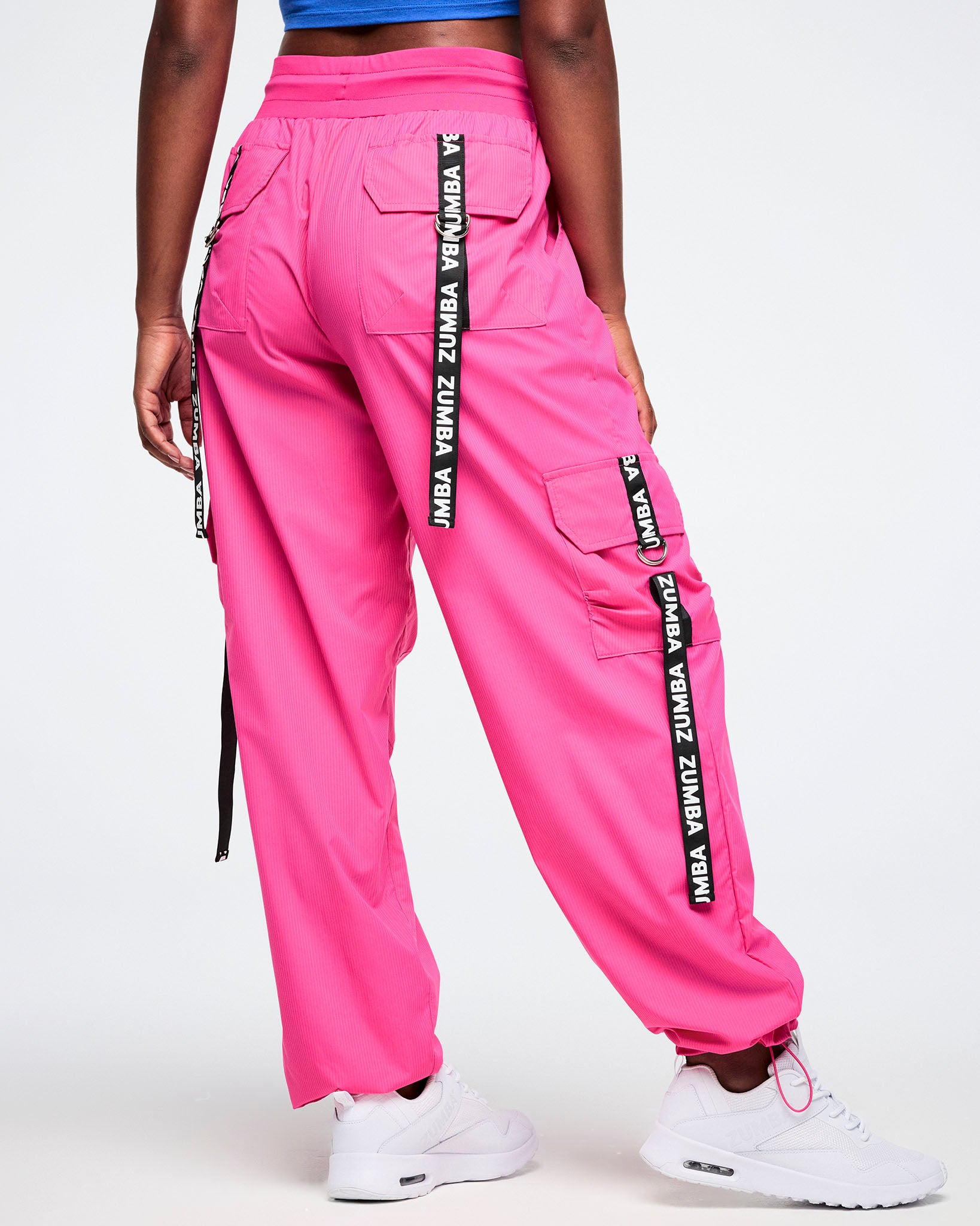 Back view of a female model wearing  bright pink cargo pants accented with black pocket ribbons featuring white Zumba lettering, styled with white shoes.