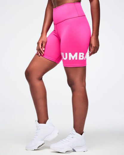 Side view of a woman wearing pink Zumba biker shorts with white Zumba branding on the left leg, styled with white shoes.
