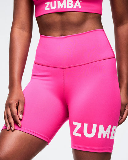 Close-up of female model wearing pink Zumba biker shorts and pink racerback with white Zumba branding on each.