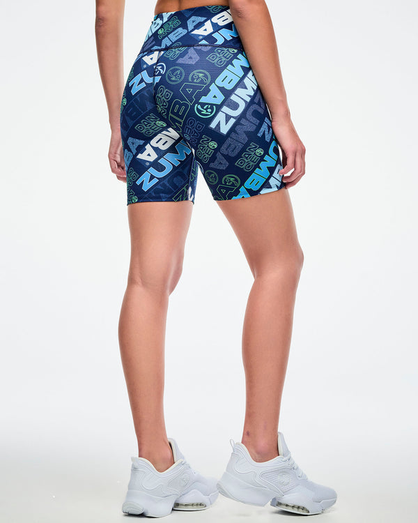 Back view of a female model wearing navy biker shorts with a blue and green “ZUMBA” logo pattern paired with white shoes.