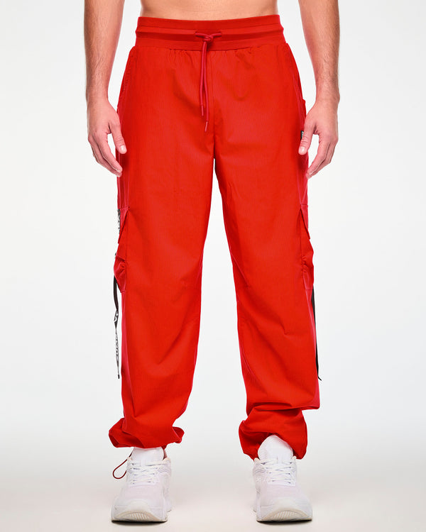 Male model in the red cargo pants with black 'ZUMBA' webbing straps on both cargo pockets, a wide waistband with a red drawcord, and red bungee cords at the ankle cuffs. Paired with white shoes.
