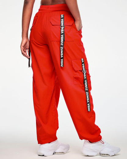 Shown from the back, a female model in the red cargo pants, with 'ZUMBA' webbing straps and silver D-rings on both the back pocket and the side cargo pocket.