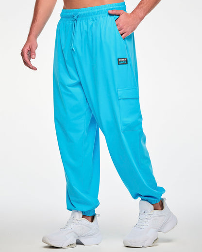 Male model in aqua blue baggy cargo pants with a loose, relaxed fit, shown in side view with a black 'ZUMBA' X 'THE SMURFS' patch on the left hip and the flap cargo pocket on the outer leg.