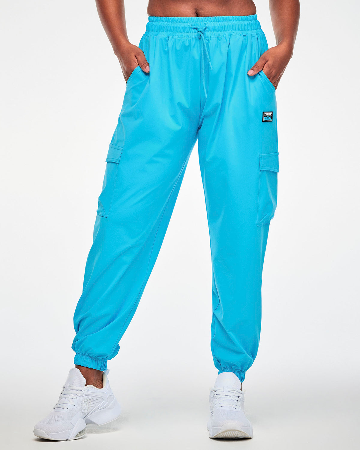Female model with both hands in the pockets of aqua blue baggy cargo pants, featuring an elastic drawcord waist, flap cargo pockets on both legs, elastic ankle cuffs, and a black patch with white 'ZUMBA' and blue 'THE SMURFS' logos on the upper left leg.