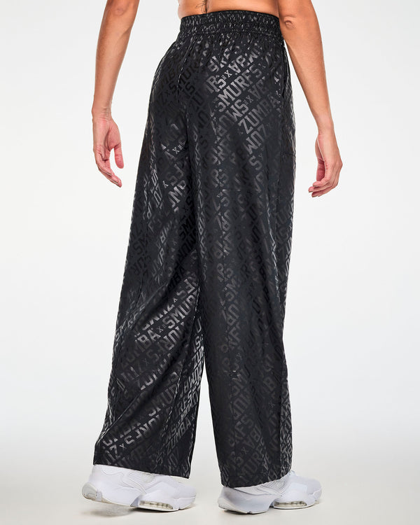 Back view of a female model in the black tonal logo print wide-leg pants, showing the diagonal 'ZUMBA' X 'THE SMURFS' print across the seat and full wide legs that fall over white shoes.