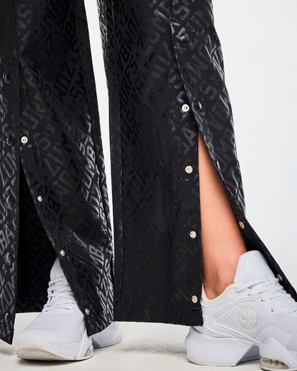 Close-up of the wide-leg pant hems with the snaps undone, showing black snaps and silver buttons along each front slit against the glossy tonal logo print.
