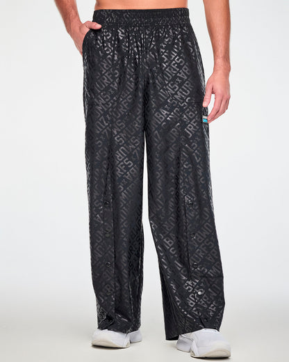 Male model with one hand in the pocket of the black tonal logo print wide-leg pants, showing the elastic waistband and black snaps down the front of each leg. Paired with white shoes.