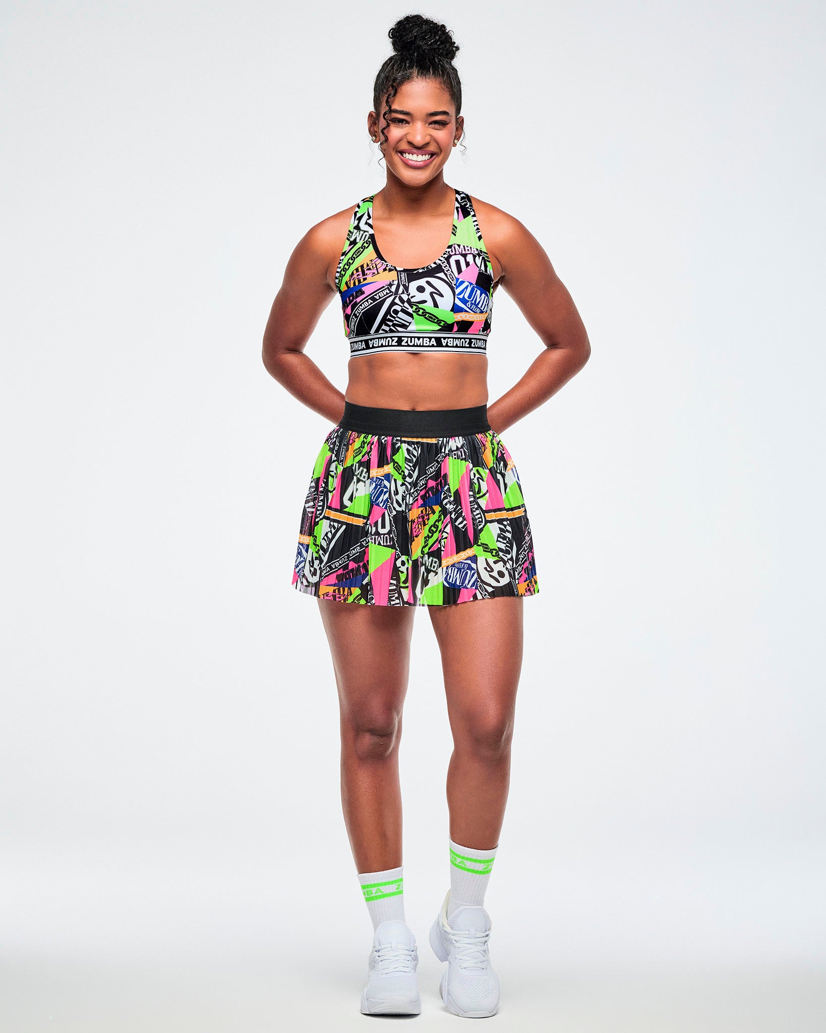 Full view of a female model wearing a skort with a vibrant graffiti-style print in neon green, pink, yellow, and black, paired with green and white socks and white sneakers.