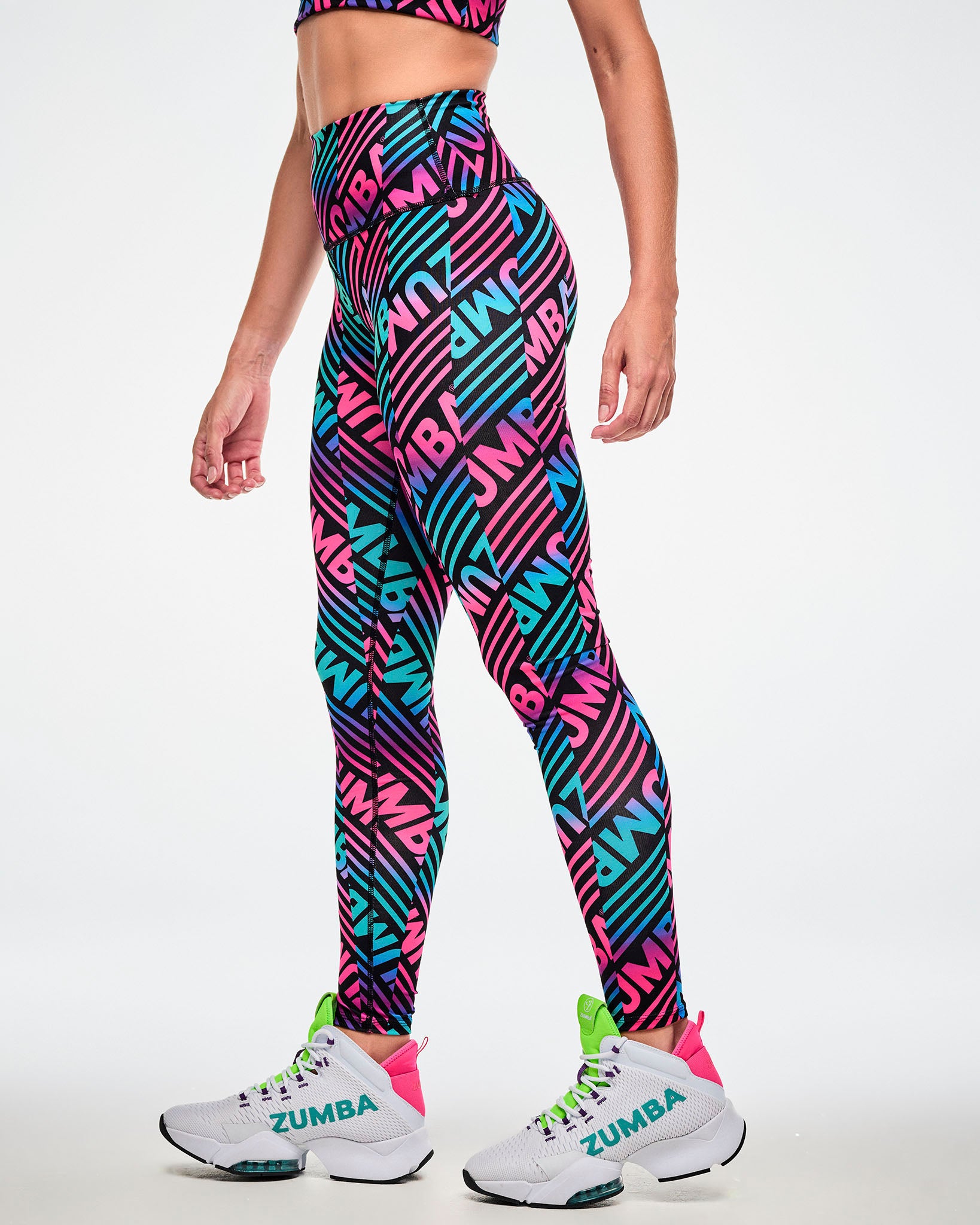 Female model wearing high-waisted ankle leggings with a repeated pink, aqua, and black "ZUMBA" print, paired with white and green shoes, side view.
