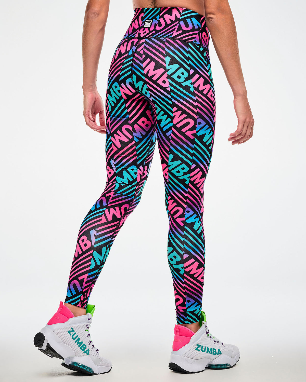 Shown from the back, female model wearing high-waisted ankle leggings with a colorful diagonal-striped and paired with white "ZUMBA" shoes.