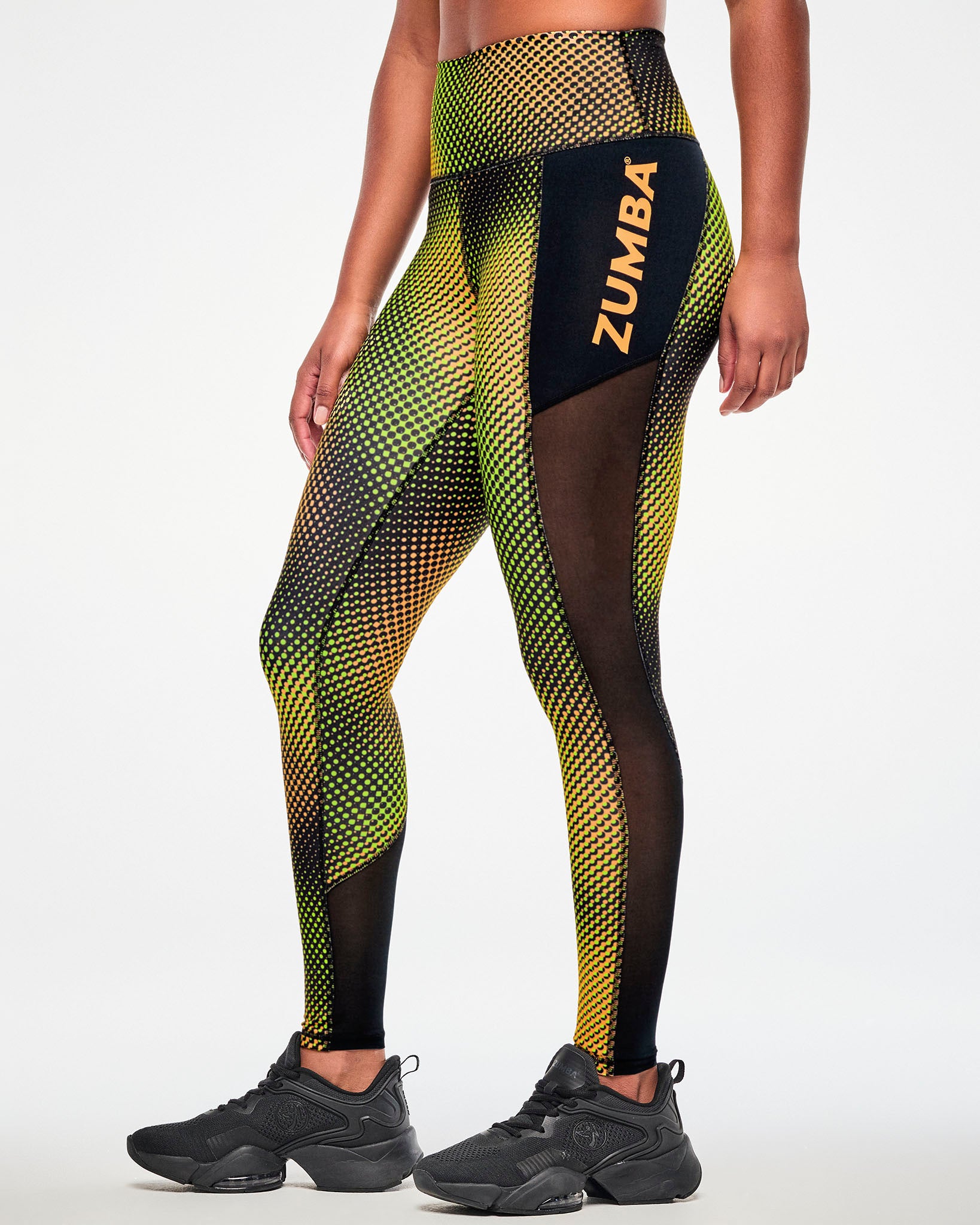 Side view of black leggings with a yellow and orange halftone dot gradient print and a mesh side panel with orange "ZUMBA" text and paired with black shoes.