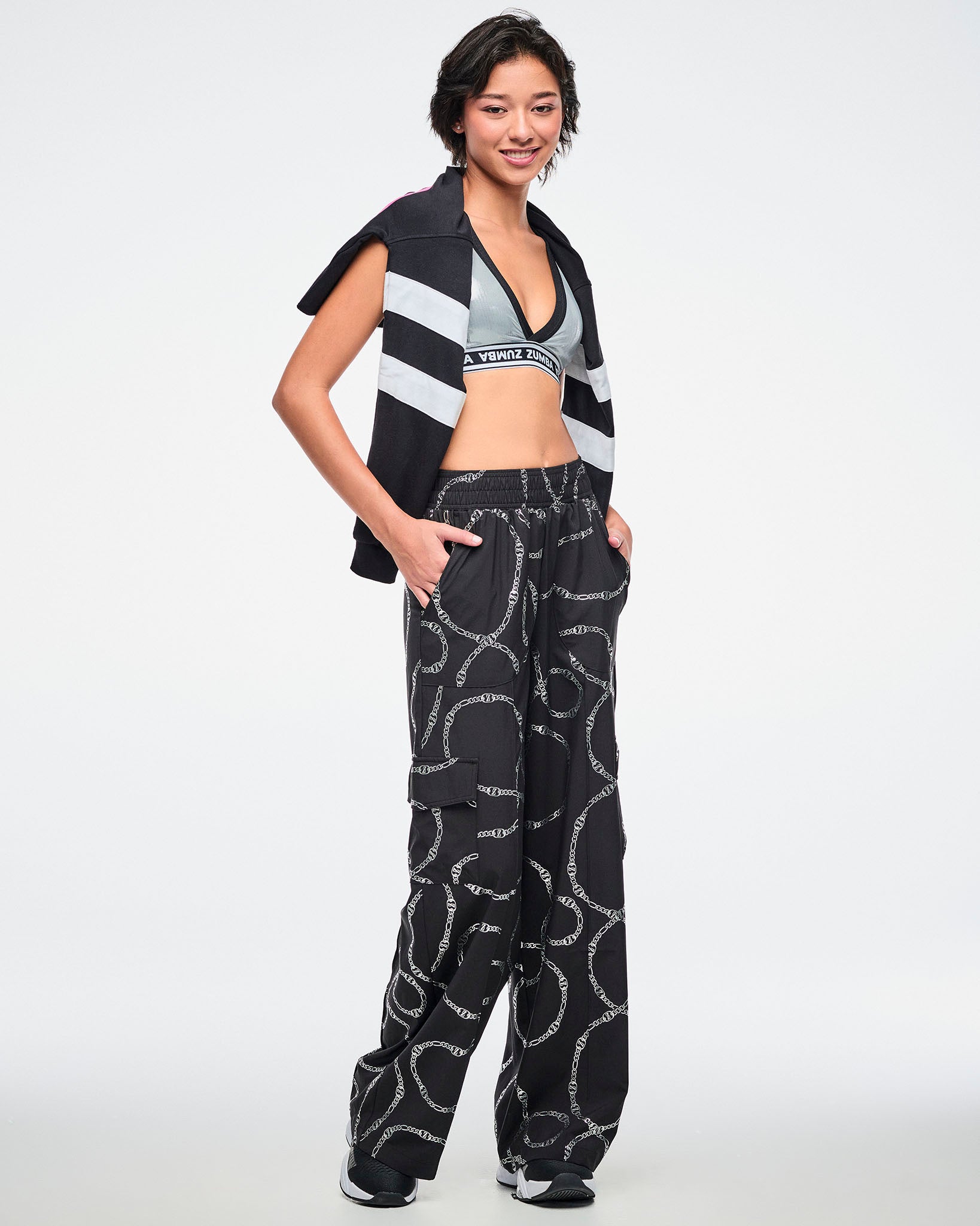 Female model wearing black and silver chain-patterned cargo pants, paired with a silver bra and a black-and-white polo draped over her shoulders.