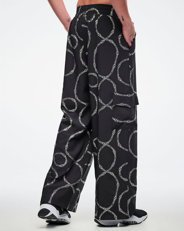 Back view of a male model wearing black cargo pants with silver chain pattern, paired with black and white shoes.