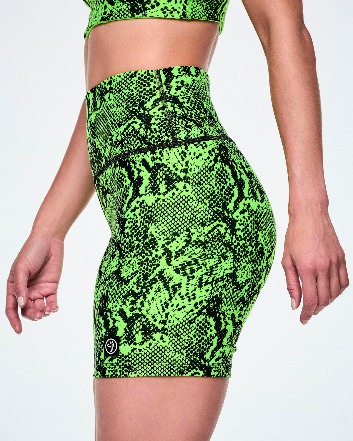 A close-up of a female model wearing neon green high-waisted biker shorts and a matching crop top with a bright neon green and black snakeskin pattern, with a small Zumbito logo near the hem.