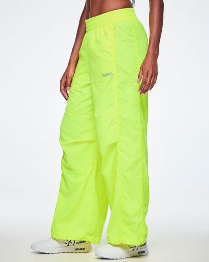 A female model wearing bright neon yellow-green wide-leg cargo pants with a small silver ZUMBA text on the left thigh, paired with white shoes.