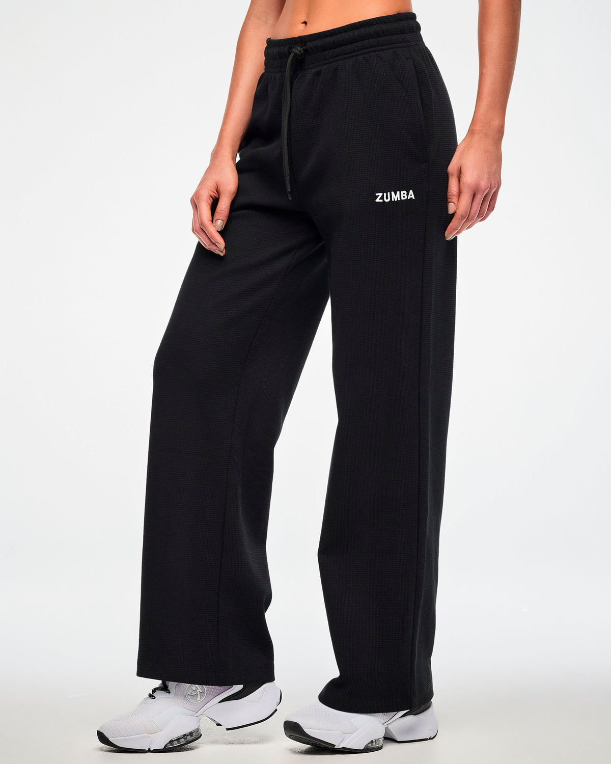 Black waffle-knit wide-leg track pants with an elastic drawstring waistband and a small white 'ZUMBA' logo on the upper left leg,  female model.