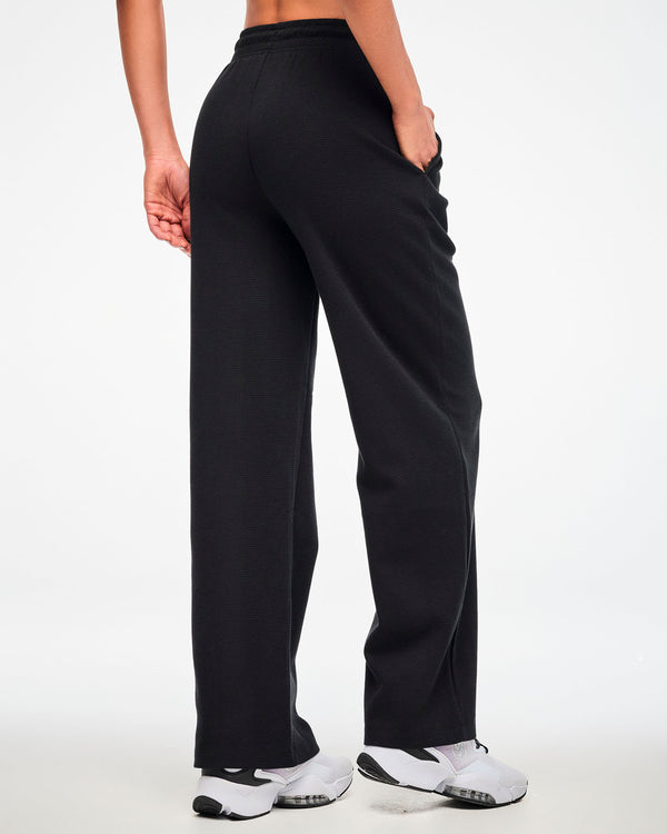 Back view of the black waffle-knit wide-leg track pants, female model with one hand in a side pocket.