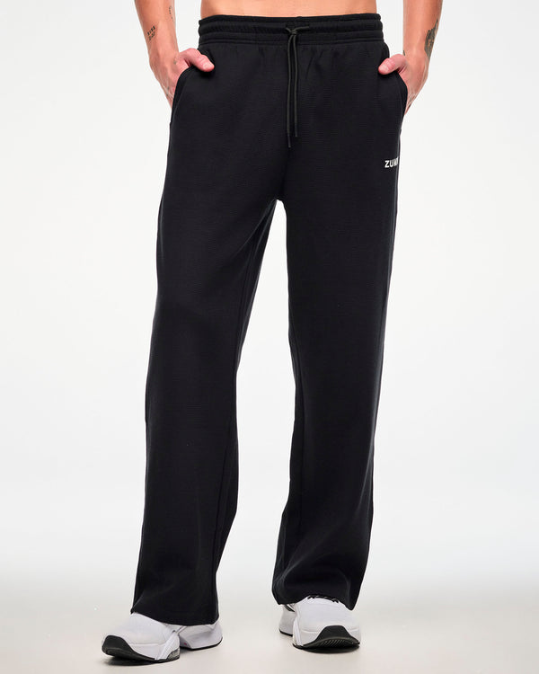Front view of a male model wearing the black waffle-knit wide-leg track pants with both hands in the side pockets.