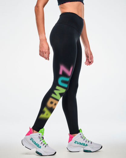 Female model wearing black high-waisted ankle leggings with a dot-gradient graphic "ZUMBA" text, in pink, aqua, green, orange and yellow text down the right leg, paired with white shoes.
