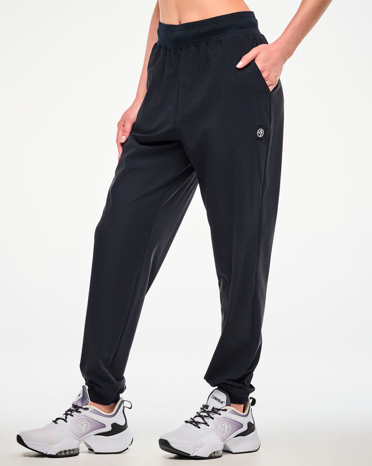 A female model wearing high waisted black jogger pants with a small white Zumbito below the left pocket and paired with white shoes.
