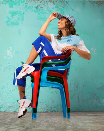 White Zumba high-top dance sneakers with red “ZUMBA” logos, green tongue, neon yellow lace details, and chunky cushioned soles. Female model wears a blue-and-white color block tee, royal blue track pants with white side stripes, and a blue patterned cap, lounging on stacked red, green, and blue chairs against a teal wall.