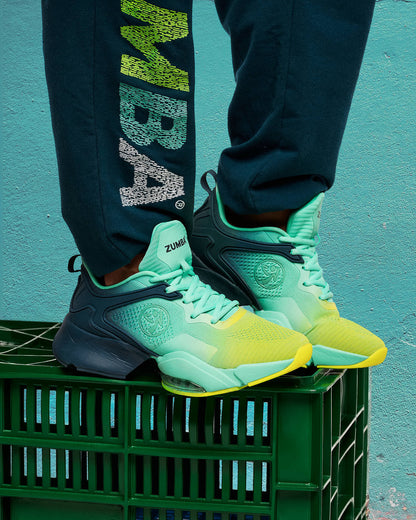 Teal-to-yellow gradient Zumba low-top dance sneakers with mint laces, molded navy heel cage, and chunky sole with clear air unit. Model wears dark teal joggers with multicolor “ZUMBA” text down the leg, standing on a dark green plastic crate against a teal wall.