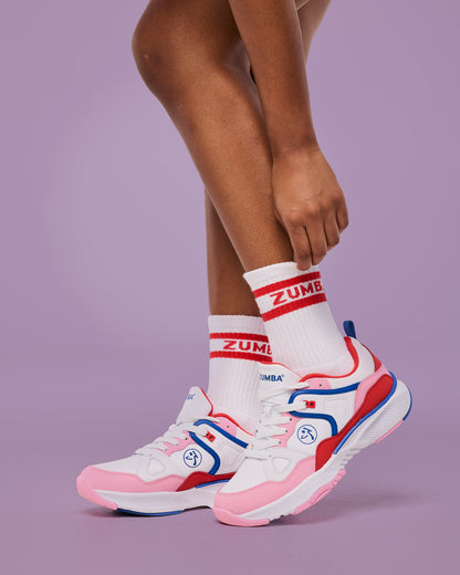 Female model wearing white crew socks with red stripes and “ZUMBA” text, paired with white athletic sneakers accented in pink, red, and blue with a Zumba logo on the side, posed against a lavender studio backdrop.